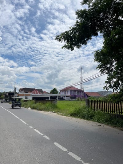 The image depicts a road scene in the city of Langsa, Indonesia. In the foreground, a paved road with a dashed white line runs diagonally across the frame from the bottom left to the upper right. A three-wheeled vehicle, resembling a motorized tricycle, is traveling away from the viewer on the left side of the road. Beyond the road, a patch of tall green grass lines the roadside, interspersed with scattered debris. A dark-colored metal fence runs along the edge of the grassy area.

In the middle ground, several buildings are visible. To the left, a long, low-rise structure with a dark roof and signage that includes the words "GARASI BAGUS" is present. Further to the right, a distinct two-story house painted purple with white trim stands out, featuring balconies and a red-tiled roof on an adjacent section. Behind these houses, a tall cell phone tower is prominent against the sky. Power lines are strung across the scene, supported by utility poles.

The background is dominated by a bright blue sky filled with scattered, textured white clouds. In the upper right corner, the dark green foliage of a large tree partially frames the scene. The overall impression is a daytime view of a suburban or semi-rural area.