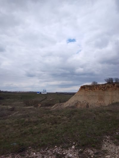 This image captures a wide, expansive rural landscape under an overcast sky. In the foreground, the ground is covered with dry, sparse grass and scattered small white and grey rocks. Moving towards the midground, the terrain features undulating hills and depressions, with more patches of dry grass and some bare bushes or small trees, suggesting an early spring or late autumn setting.

A prominent feature on the right side of the midground is a steep, erosion-formed cliff or earth cut, revealing layers of light brown and reddish-brown soil and rock formations. The top of this cliff is relatively flat, with a few bare, shrub-like trees.

In the distance, towards the left of the frame, stands a white building with distinctive blue dome-shaped roofs and spires, resembling a church or monastery. A faint dirt path appears to lead towards this structure across the rolling hills. A line of bare trees forms a distant horizon behind the church, separating the fields from what might be a forested area or more distant terrain.

The sky occupies a significant portion of the upper frame, dominated by heavy, grey clouds. A small, distinct patch of blue sky is visible roughly in the upper-middle section, offering a slight contrast to the otherwise cloudy expanse. The lighting suggests a cloudy daytime, possibly late morning or afternoon. There are no people or discernible activities taking place. No text is visible in the image. The scene depicts a quiet, natural landscape, characteristic of a rural area in Yablonovo, Russia.