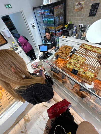This image depicts the interior of a commercial establishment, likely a bakery or snack shop, located in Asnières-sur-Seine, France. In the immediate foreground, partially visible from behind, a person with long blonde hair wears a dark, possibly sequined jacket. This individual holds a smartphone in their left hand, oriented as if capturing an image or video. A red handbag rests beside their right leg, which displays a tattoo. Behind a glass display counter, a female employee with dark hair pulled back, wearing a black top with a logo, stands at a point-of-sale terminal, which features a blue screen. To her right, a tall, glass-fronted refrigerator is stocked with various bottled and canned beverages. The counter behind her is white and includes a microwave oven and beverage supplies. The display counter showcases a large basket of golden-brown, glazed pastries and multiple rectangular flatbreads, resembling pizza slices, arranged on wooden stands with price labels, including one indicating "2,50 €". Below the visible food, further pastries are displayed within the glass case. In the background, through an open doorway, a second employee is partially visible, wearing a dark garment and a purple apron, positioned near a sink area. The establishment features light-colored, square wall tiles and a patterned marble floor. An emergency exit sign is affixed above the doorway.