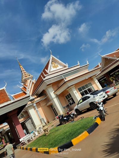 The image shows a sunny daytime scene in Bavet Quarter, Cambodia. A modern building with ornate, traditional Khmer architectural elements, including multi-tiered roofs with decorative finials and gable ornamentation, is prominently featured. The building has peach-colored walls and large windows. In the foreground, there are several parked motorcycles and two cars, one white SUV and one silver sedan. A patch of green grass with a black and yellow border is visible. Two individuals are partially visible on the right side of the image, near the cars. Another individual in a green shirt and grey pants is walking away from the camera on the left side of the frame. The sky is blue with scattered white clouds. A watermark indicating "realme Shot by zsm_travel 2026.06.24 09:17" is present in the lower right corner. The scene appears to be an area for vehicle processing or a public thoroughfare associated with the building.
