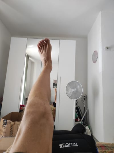 The image shows a first-person perspective of a person's leg and foot raised in the air. The person is indoors, likely in a bedroom, given the presence of a large mirrored wardrobe, a fan, a ceiling light fixture, and a box that appears to be a monitor or television box. The leg is bare and shows some light hair. The foot is positioned upwards, with toes clearly visible. The background includes a white wall, the mirrored wardrobe which reflects a portion of the room, and some cleaning tools leaning against the wall. The lighting appears to be natural, possibly from a window. The scene is domestic and unremarkable, with no indication of being in Grosseto, Italy.