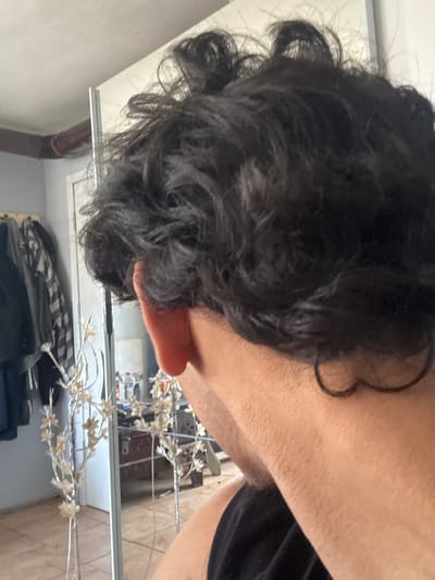 The image shows a close-up view of a person's head and upper body reflected in a mirror. The person has dark, curly hair and fair skin, visible around their ear and neck. They are wearing a black t-shirt. The background reflection shows a cluttered room with what appears to be a decorative, silver, star-shaped light fixture. Part of a wardrobe or closet is visible near the bottom of the reflection. It is not possible to provide the location in Novi Pazar, Bulgaria from this image alone.