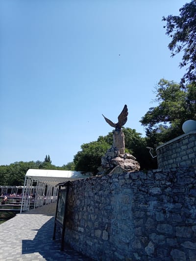 A low-angle, wide shot captures an outdoor scene with a prominent stone sculpture of an eagle with outstretched wings, perched on a rocky base. The sculpture stands against a clear blue sky, with lush green trees forming a backdrop. To the left, a paved walkway with a white canopy stretches along a body of water, where several people are visible relaxing or walking. A rustic stone wall runs along the right side of the image, leading towards a rounded structure topped with a sphere. The overall setting appears to be a park or recreational area, likely in Zheleznovodsk, Russia, known for its natural beauty and spa resorts.