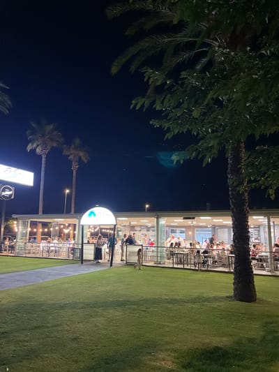 The image depicts a nighttime scene at a restaurant with a large outdoor seating area. The restaurant, identified by signage as "El Sira," is illuminated by string lights and interior lighting, showcasing diners seated at tables. Palm trees are visible in the foreground and background. The area appears to be a grassy lawn with a paved walkway leading to the restaurant. The overall setting suggests an evening meal at an establishment in Canet d'en Berenguer, Spain, possibly near a coastal area given the presence of palm trees.