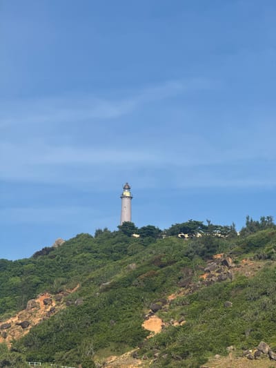 The image shows a lighthouse on a cliff overlooking the sea. The lighthouse is a tall, white structure with a black lantern room. It is surrounded by lush green vegetation and rocks. The sky is clear blue with some wispy clouds. The sun appears to be high in the sky, suggesting it is midday. The location appears to be a coastal area, possibly a tourist destination given the presence of a white railing visible at the bottom right, indicating a walkway or viewing platform. The overall scene is serene and picturesque. There is no visible text in the image.