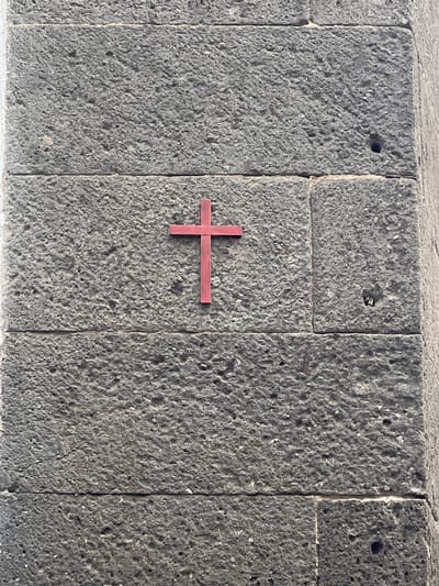 The image shows a close-up of a rough, gray stone wall, likely constructed from volcanic rock. The wall appears to be made of rectangular blocks fitted together. A red cross is affixed to the center of the wall. The wall is located in Las Palmas, Spain. There are no people visible in the image.