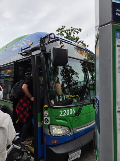 A green and blue RapidBus is stopped at a transit stop in West Vancouver, Canada. The bus, numbered B22006, has its doors open and passengers are boarding. A man in a black t-shirt and red and black plaid shirt is stepping onto the bus, carrying a backpack. Another person, mostly obscured, is behind him. In the foreground, a person in a grey hoodie is visible pushing a stroller. A bus stop sign is on the right side of the image. The sky is overcast.