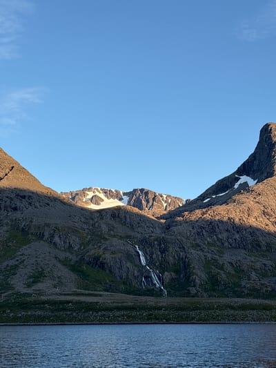 The image depicts a scenic view of the coast in Arnøyhamn, Norway, with dramatic mountains meeting a calm body of water. The sky is clear and blue with a few wispy clouds on the left. The mountains are rugged and rocky, with patches of snow visible on the higher peaks. A waterfall cascades down the mountainside in the middle ground. The foreground shows a body of water, likely the sea, with gentle ripples on its surface. The lighting suggests it is either early morning or late afternoon, with the sun casting long shadows across the mountains. There are no people or man-made structures immediately visible, emphasizing the natural beauty of the landscape.