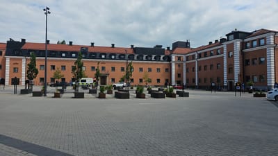 The image depicts a large, brick building with a red tiled roof, located in Linköping, Sweden. The building appears to be a government or administrative facility, with multiple windows and entrances. In the foreground, there is a paved courtyard with several planter boxes and seating areas. A few cars and bicycles are visible in the background, as well as a few people. The sky is overcast, suggesting a cloudy day. The architecture of the building is traditional, with symmetrical elements and decorative trim. The overall scene conveys a sense of order and public space.