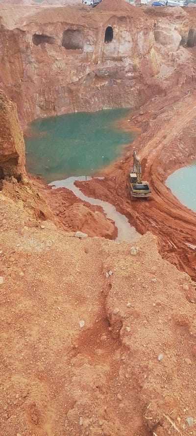 A large-scale excavation is underway in an open-pit mine. The terrain consists of reddish-brown earth, with steep, layered walls revealing the strata of the soil. Several pools of murky, turquoise water have accumulated at the bottom of the pit, indicating water infiltration.
In the middle ground, a yellow excavator with the "CAT" logo is positioned on a muddy track, facing away from the viewer. It appears to be actively engaged in or recently finished moving earth. Another smaller body of water is visible to the right of the excavator.
The background shows the upper rim of the pit, where more disturbed earth and some vehicles, including what appear to be trucks and a blue car, are present. The overall scene suggests ongoing construction or resource extraction. The location is described as the city of Reifild-Ist, Nigeria.