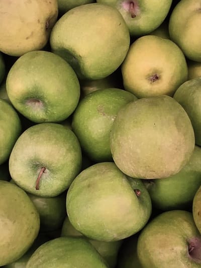 A close-up, top-down shot displays a bountiful pile of green apples. The apples are various shades of green, with some exhibiting yellow undertones and subtle reddish blush marks. The smooth, round surfaces of the fruits create a textured pattern, with their stems visible on some. The lighting is even, highlighting the organic shapes and soft textures of the apples, suggesting they are fresh and ripe. This image could be from a local market or orchard in Voluntari, Romania, showcasing a common agricultural product.