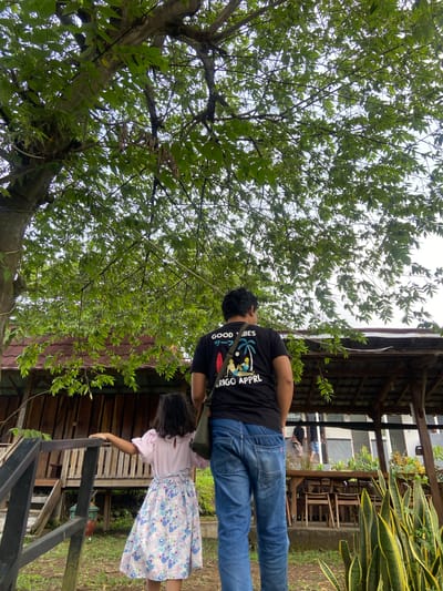 A man in a black T-shirt, blue jeans, and a green shoulder bag stands near a girl wearing a light pink shirt and a floral dress. They are both positioned near a wooden structure, likely a porch or elevated walkway. The surrounding environment is lush with green foliage, including a large tree with dense leaves. The scene takes place outdoors, and the location is Penanggungan, Indonesia.