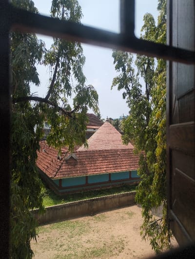 The image displays an outdoor scene viewed through a dark-framed window, with partial wooden shutter elements visible on the right. A traditional Kerala-style building with a multi-layered, red-tiled roof dominates the middle ground. The roof features a central, elevated pyramid-shaped section surrounded by sloping tiled surfaces. The building's exterior walls are painted a distinct light teal or turquoise color, with vertical decorative elements. Immediately surrounding the building is a narrow strip of green grass, separated by a low concrete wall from a larger foreground area of dry, light brown earth with sparse green patches. Dense green trees with abundant foliage frame the building on both sides and extend upwards into a clear, light blue sky. No individuals or animals are present. The architectural style is consistent with traditional structures found in Kochi, India.