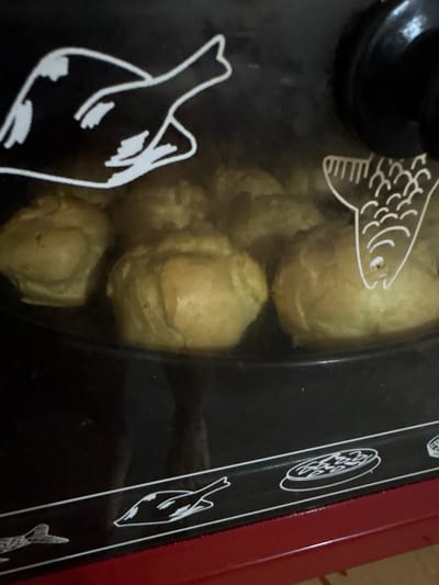 The image shows a close-up view of a red and black appliance, likely a small oven or grill, with a glass lid. Inside the appliance, several golden-brown, round food items are visible, possibly pastries or baked goods. The glass lid has white line drawings on it, including a stylized fish on the left and a bird-like figure on the right, with more decorative circular patterns in between. The appliance appears to be in a domestic setting, with hints of a wooden surface or floor visible in the background. The lighting suggests it is daytime, but no specific time of day can be determined. There is no visible text in the image.