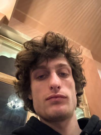 A low-angle selfie reveals a young man with curly brown hair and stubble. His blue eyes gaze towards the camera. He's wearing a dark hooded sweatshirt. The background is a pale orange wall, and a window with a gold frame is visible. A reflection, possibly a chandelier, is seen in the window. The perspective makes him appear to be looking down at the viewer.