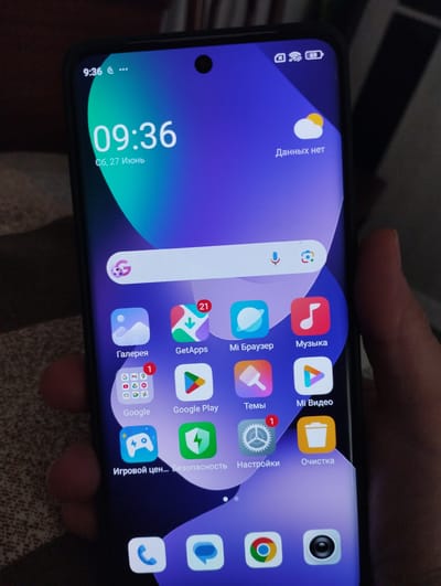 This is a close-up, eye-level photograph of a smartphone screen displaying a colorful wallpaper and various app icons. The phone is held in a person's hand, partially visible on the right side of the frame. The time on the phone reads "09:36" and the date is "Сб, 27 Июнь," which translates to "Sat, June 27". The battery indicator shows 69%. Visible app icons include "Галерея" (Gallery), "GetApps", "Mi Browser", "Music", "Google Play", "Themes", "Mi Video", "Game Center...", "Security", "Settings", and "Cleaner". The context of Yangiiul, Uzbekistan is not present in the image.