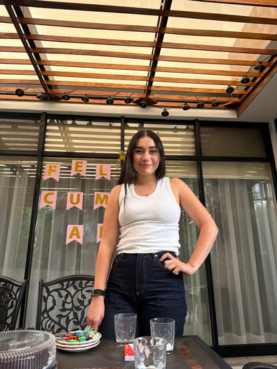 The image shows a woman standing at a table on a patio or balcony. She is wearing a white tank top and dark jeans. She is smiling and looking at the camera. On the table, there is a decorated cake, glasses, and other items. In the background, there is a window with a "FELIZ CUMPLE" banner hanging on it. The patio is enclosed by a wooden structure with lights. The location is Barrio de Nuevo León, Mexico.