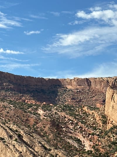 The image displays a vast, arid mountainous landscape under a partly cloudy blue sky. The terrain is characterized by steep, rugged slopes covered in reddish-brown rock formations, scattered boulders, and sparse, low-lying green vegetation. A prominent, layered cliff face dominates the upper portion of the image, exhibiting distinct horizontal striations. The overall impression is one of a remote, natural environment, likely a canyon or desert region, with no signs of human habitation or activity.