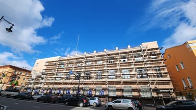 The image shows a street in Smolensk, Russia. A multi-story building with scaffolding covering its facade is the main subject. The building is light beige in color with a visible antenna on top. Below the scaffolding, there is a black metal fence with informational posters. Cars are parked along the street in front of the building and fence. A streetlamp is visible on the left side of the frame. The sky is blue with white clouds. Other buildings with yellow or orange facades are visible to the sides and left of the primary building. The weather is sunny and clear.