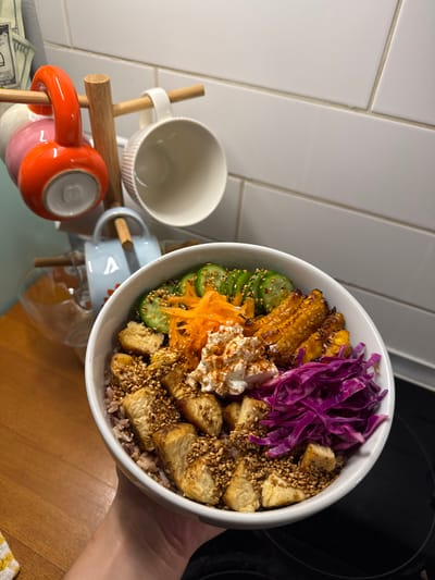 A person holds a bowl filled with various foods, including sliced cucumbers sprinkled with sesame seeds, grilled corn, shredded purple cabbage, shredded carrots, a dollop of cream, and tofu cubes with sesame seeds. Beneath the ingredients, there appears to be rice. The bowl is held over a wooden surface. In the background, there are ceramic mugs on a wooden stand and white tiled walls.