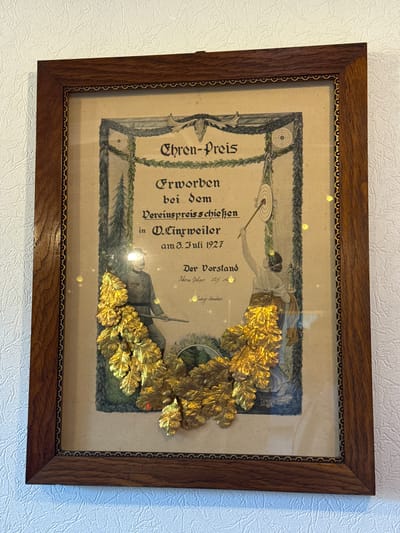 The image shows a framed certificate hanging on a textured white wall. The certificate is ornate, with a dark wooden frame featuring decorative borders. The certificate itself is light brown and features a central illustration. The illustration depicts two figures: on the left, a man in a military-style uniform holds a rifle, and on the right, a woman in a classical gown aims an arrow at a target. A garland of golden oak leaves is prominently placed across the bottom of the illustration, overlapping the figures. The text on the certificate is in German and appears to be an award or recognition. Some of the legible text includes "EHREN-PREIS", "Erworben bei dem Vereinspreisschießen in O. Eingweiler am 3. Juli 1927", and "Der Vorstand". Based on the text, this certificate was awarded at the shooting competition in O. Eingweiler (likely Oberlinxweiler), a district of Sankt Wendel, Germany, on July 3, 1927. The overall scene is well-lit, with some reflections visible on the glass covering the certificate.