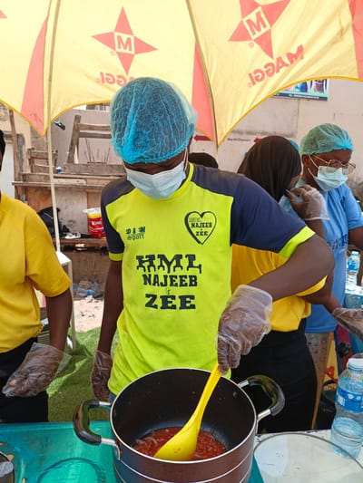 The image depicts an outdoor scene in Tarauni, Nigeria, where individuals are preparing food. A person in the foreground, wearing a blue hairnet and a light blue surgical mask, is stirring a pot of red sauce with a yellow ladle. They are also wearing a bright yellow and dark blue t-shirt with the text "HML NAJEEB & ZEE" and the "SPORT" logo. They are also wearing clear plastic gloves, as are the other individuals visible. To the left, another person in a yellow t-shirt is partially visible. In the background, under a yellow umbrella with a red star logo and the word "MAGGI" printed on it, other individuals can be seen. One person is wearing a black hijab and a blue t-shirt, while another to their right wears a light blue hairnet, glasses, a surgical mask, and a light blue t-shirt. Various items are visible in the background, including a white bucket with a red label, plastic bottles of water, and what appears to be a food stall setup. The environment suggests a busy marketplace or outdoor event.