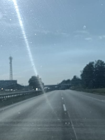 The image depicts a view from inside a vehicle, looking through a dirty windshield at a highway during daytime. The sky is overcast and appears a muted blue-grey, suggesting cloudy weather. The highway stretches into the distance, with a white line marking the center. To the left of the highway, a railing or barrier is visible, and beyond that, a dark building with a tall, lattice tower structure is present. Further to the left, there are also some blurred lights which could be from other vehicles or infrastructure. To the right of the highway, silhouetted trees form a dark, dense line against the sky. The overall impression is that of a journey on a highway in a somewhat drab or subdued environment, possibly on a day with poor visibility or low light conditions. The streaks and dust on the windshield are prominent, affecting the clarity of the scene and adding a textured, slightly hazy quality to the image. There is no visible text in the image. The scene conveys a sense of travel and transit.