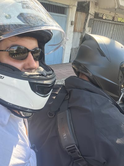Two individuals are wearing helmets and riding a motorcycle in the city of Buleleng, Indonesia. The person in the foreground is wearing a white and black helmet with a clear visor, sunglasses, and a white shirt. The person in the background is wearing a black helmet and a black jacket, with a dark backpack strapped on. In the background, there is a building with blue doors and windows, and a sign that reads "TOKO GADING SEMPURNA". The scene appears to be during the daytime, with sunlight casting shadows.
