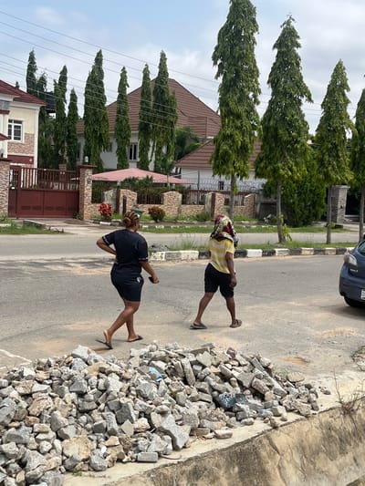 Two women are walking on a paved street in Abuja, Nigeria, near residential buildings. The woman on the left is wearing a black t-shirt and black shorts. Her hair is styled in braids. The woman on the right is wearing a yellow and white striped t-shirt and black shorts, with a patterned fabric covering her head. She is holding her right hand to her head. In the foreground, there is a pile of grey rubble and construction debris next to a concrete drainage channel. Behind the women, there are several tall, slender green trees lining the street, with houses and fences visible in the background. Power lines are strung overhead. A dark-colored car is parked on the right side of the road. The sky is overcast.