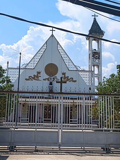 The image depicts the facade of a white church in Diên Khánh, Vietnam, partially obscured by overhead power lines and a large metal gate in the foreground. The church features a prominent triangular gable with intricate white decorative cutouts, a large circular golden emblem, and golden dragon motifs. A statue of a saint stands to the right of the emblem, next to a large cross. To the right of the main structure, a white bell tower with a dark green, tiered roof rises against a blue sky with fluffy white clouds. The bell tower also has decorative cutouts and a cross at its apex. Green trees are visible on either side of the church building. The white metal gate in the foreground is adorned with golden scrollwork, and behind it, the entrance to the church grounds features more decorative metalwork and a covered walkway. The ground in front of the gate is a paved surface, casting shadows from the gate and nearby objects.