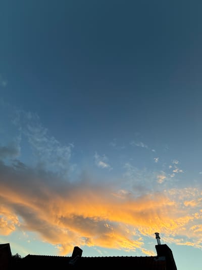 The image depicts a sunset viewed from a rooftop in Bruges, Belgium. The sky is a deep blue, transitioning to lighter shades towards the horizon, with scattered clouds illuminated by the setting sun in hues of orange and yellow. Silhouetted against the sky are the dark rooftops of buildings, including a chimney with a metal cap. The scene is serene and devoid of any visible people or activity. The architecture suggests a European city setting.