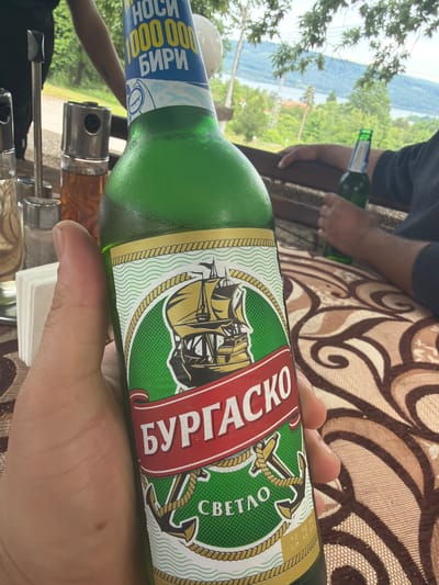The image, taken in Sushina, Bulgaria, captures a serene outdoor scene centered around a person holding a green beer bottle. The foreground is dominated by a hand gripping a bottle of "Burgasko" beer. Its label features a sailing ship, anchors, and rope details, with prominent Cyrillic text reading "БУРГАСКО" (Burgasko) and "СВЕТЛО" (Svetlo), indicating a light beer. A blue and white neck label further states "НОСИ 100 000 000 БИРИ" (Nosi 100,000,000 Biri), a promotional slogan, along with small numbers "12 10 23" possibly indicating a date.

In the mid-ground, part of another person, seated at a table, is visible, holding a second green bottle. The table is covered with a patterned brown and cream tablecloth, and metal condiment dispensers are also partially visible.

The background reveals a picturesque daytime landscape from a high vantage point. A vast, calm body of water, likely a river or lake, stretches across the mid-distance, surrounded by lush green, tree-covered hills or mountains under a clear sky. The setting suggests a relaxing moment, possibly at an outdoor cafe or restaurant, enjoying drinks with a scenic view in good weather.