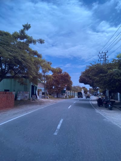 An overcast day in Aluepiet, Indonesia, presents a view down a two-lane asphalt road. Trees with lush green foliage and some with autumnal brown leaves line the road, interspersed with buildings. On the left, a light green building is visible with stacks of red bricks and what appear to be tiles or slabs of material in front. A dirt path runs alongside the road. On the right side of the road, several parked motorcycles are visible, along with a utility pole carrying power lines. Further down the road, two vehicles are in motion: a dark truck on the left lane and a smaller cart-like vehicle in the right lane. A decorative fence with white posts and green accents stretches along the right side of the road in the distance. The sky is a mix of blue and white clouds.