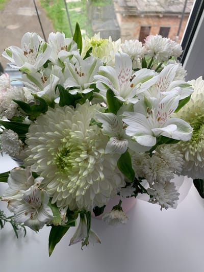 A bouquet of white and green flowers, including large chrysanthemums and smaller alstroemeria, is displayed in a pink vase. The bouquet is positioned on a white surface, likely a windowsill, with a blurred background showing greenery and a brick building, indicative of an urban environment. The scene suggests a domestic setting, possibly in Volchansk, Russia, given the architectural style visible in the background. No people are present in the image.