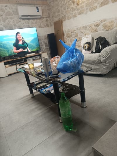 The image depicts an indoor scene in what appears to be a living room. The walls are constructed of light-colored stone. A television showing a woman is on in the background, placed on a low TV stand. A black speaker stands beside the television.

In the foreground, there is a two-tiered black glass coffee table. On the table, there are several items including a glass of beer, a partially consumed can of "Пиво Бенара" (Pivo Benara) beer, various remote controls, a plastic bag filled with items, and a pack of cigarettes. On the lower tier of the table, there are more items, including a blue package of some sort. Underneath the table sits a partially emptied green plastic bottle of carbonated water.

To the right of the coffee table is a light-colored patterned couch. On the couch, there is a shopping bag with a gold and black logo and a black backpack. 

Based on the beer can, the location is potentially in or near Bulgaria, however, the specific city of Izbeglii cannot be confirmed from the contents of the image.