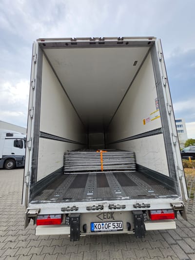 The image shows the rear interior of an open trailer truck parked on a paved lot. Inside the trailer, several stacks of what appear to be metal scaffolding or similar equipment are secured with an orange strap. The trailer walls are white with black tie-down rails. A white semi-truck is partially visible to the left of the trailer. The rear of the trailer features red taillights, a license plate reading "KO OF 530", and other mechanical components. A building and trees are visible in the background. The sky is overcast with clouds. The location is identified as Zeppelinheim, Germany.
