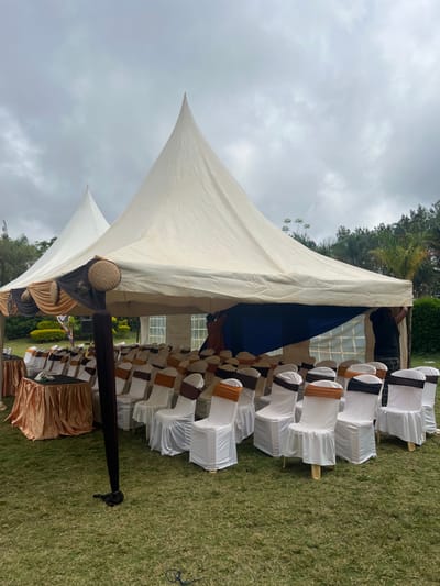 This image captures an outdoor event setup on a well-maintained grassy lawn in Makuyu, Kenya, under an overcast sky. The primary subjects are two large, light-colored pyramid-shaped tents, or marquees, with distinct pointed tops. The larger tent, positioned prominently, features a blue fabric lining or drape visible inside.
Beneath the main tent, numerous chairs are neatly arranged in rows, suggesting seating for a gathering. Each chair is covered in white fabric and adorned with sashes or ribbons in alternating colors of brown, black, and a golden or orange shade. Several tables are also visible, draped with gold or brown table skirts. Decorative elements, including woven, circular objects resembling wicker and dark fabric drapes, are strategically placed along the tent's supports. A person is partially visible near the top of the larger tent, appearing to be involved in its setup or adjustment.
The setting is a natural outdoor environment, characterized by expansive green grass and lush trees and foliage in the background. The sky is uniformly gray and cloudy, indicating an overcast day with diffused lighting. The overall scene suggests that an organized event, possibly a wedding, celebration, or formal gathering, is either being set up or is ready to commence. No discernible text is visible in the image.