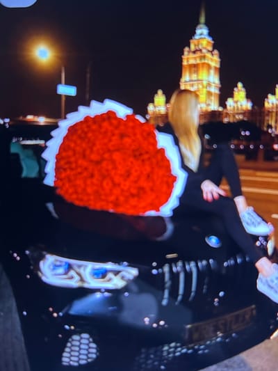 The image shows a person sitting on the hood of a black car at night. The person has long blonde hair and is wearing black clothing and white sneakers. They are sitting with their legs extended, and their left hand is resting on their right knee. In front of the person is a large, heart-shaped bouquet of red flowers with white accents. The car has a BMW logo on the hood. In the background, there are illuminated buildings, suggesting an urban setting. The streetlights and the lights from the buildings create a bokeh effect. The image is grainy and appears to be taken from a screen. The location is not Beșalma, Moldova, but rather a city with prominent, illuminated architecture.