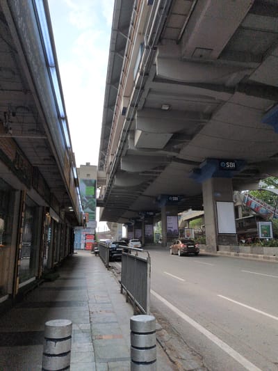 The image depicts a street-level view in an urban environment, likely in Hyderabad, India, looking forward beneath an elevated concrete structure. The scene is illuminated by natural daylight.

On the left, a paved sidewalk extends into the distance, bordered by several grey concrete bollards in the foreground. Adjacent to the sidewalk is a series of commercial establishments with glass storefronts, operating under a continuous canopy that houses fluorescent tube lights. Visible signage includes "S-160 PHOTOS XEROX" and mentions of "VISA" services.

To the right of the sidewalk, an asphalt road with visible white lane markings stretches forward. A brown sedan (Maruti Ciaz) is observed driving away in the right lane. Guardrails separate the sidewalk from the road in the mid-ground.

A large, multi-level elevated concrete structure dominates the upper and central portions of the image. This structure, characteristic of a metro line or flyover, is supported by numerous concrete pillars. Several pillars feature blue signage displaying the "SBI" (State Bank of India) logo. One pillar is marked with "C-1703" and has a blank white billboard attached. The underside of the elevated structure reveals a complex network of concrete beams and slabs. In the far right background, an escalator ascends towards what appears to be an elevated platform, consistent with a metro station. Additional elevated road infrastructure is visible further in the background. Sparse green foliage is present along the right side of the road beneath the elevated structure. No discernible people are prominently featured or engaged in specific actions.