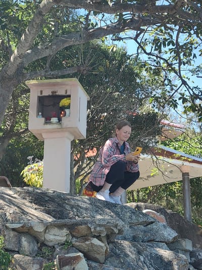 A woman crouches on a rock, engrossed in her yellow phone. She wears a pink plaid shirt over black leggings and white sneakers. Behind her stands a small white shrine adorned with flowers and incense. The background includes lush green trees, a clear blue sky, and a glimpse of a white pavilion with a red-tiled roof. The scene is bathed in bright sunlight.