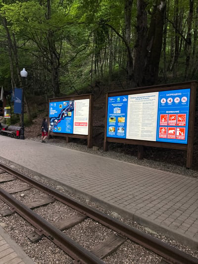The image shows an outdoor information kiosk next to a narrow-gauge railway track in a forested area. The kiosk has two large signs with information about attractions and rules. The upper sign displays safety warnings, including icons for falling rocks, slippery ice, and dangerous animals, and prohibitions like cycling and smoking. The lower sign is a map-like diagram of an excursion route within the Guamka Gorge, with accompanying images and text in Russian.

A person is visible standing near the lower sign, looking at it, dressed in a dark jacket and red shorts. The setting appears to be a nature park or scenic route, with dense green trees and foliage dominating the background. The railway tracks are made of steel rails and wooden sleepers, with gravel ballast. The pathway next to the tracks is paved with interlocking concrete blocks. A lamppost with a white globe and a bench are also present. The lighting suggests it could be daytime, possibly overcast or late afternoon, given the soft illumination and shadows. The overall atmosphere is calm and informative, aiming to guide visitors through the natural attractions while emphasizing safety precautions. The visible text is in Russian, with titles like "КАРТА-СХЕМА ЭКСКУРСИОННЫХ ДОСТОПРИМЕЧАТЕЛЬНОСТЕЙ ГУАМСКОГО УЩЕЛЬЯ" (Map-Scheme of Excursion Attractions of the Guamka Gorge) and "ПРАВИЛА ПОСЕЩЕНИЯ АТТРАКЦИОНА «ПОЕЗД НА РЕЛЬСАХ»" (Rules for Visiting the Attraction "Train on Rails").