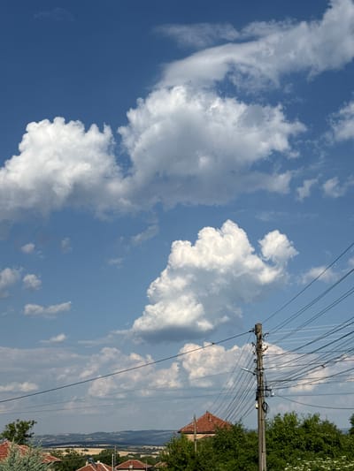 The image displays a scenic view of Slavyani, Bulgaria, under a bright blue sky dotted with fluffy white clouds. In the foreground, a weathered wooden utility pole stands tall, intricately tangled with a web of electrical wires. The pole is equipped with a street lamp and various electrical components. Behind the pole, a cluster of lush green trees and the terracotta rooftops of houses peek through, suggesting a residential area. In the distance, rolling hills create a picturesque landscape, partially obscured by a haze. The overall scene captures the serene beauty of a Bulgarian countryside, with a touch of urban infrastructure.