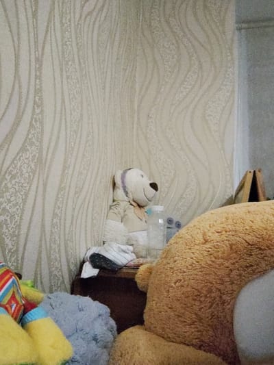 The image depicts an indoor scene, likely a bedroom or play area, with a wall covered in patterned wallpaper. In the foreground, a large, fuzzy, tan-colored stuffed bear is partially visible, occupying the right and lower portions of the frame. To the left of this bear, a smaller, white stuffed bear with a purple ribbon around its neck sits upright. It is leaning against a dark brown wooden surface, possibly a dresser or shelf. Next to the white bear, there is a clear plastic bottle and some folded clothing items, including a striped sock. Further to the left, the corner of a colorful stuffed toy with stripes and diamond patterns is visible, along with a fuzzy, light blue stuffed animal. In the background, the wallpaper has a wavy, organic pattern. The scene is lit by artificial light, creating a slightly dim atmosphere. There is no indication of the city of Okhtyrka, Ukraine, in the visual information provided.
