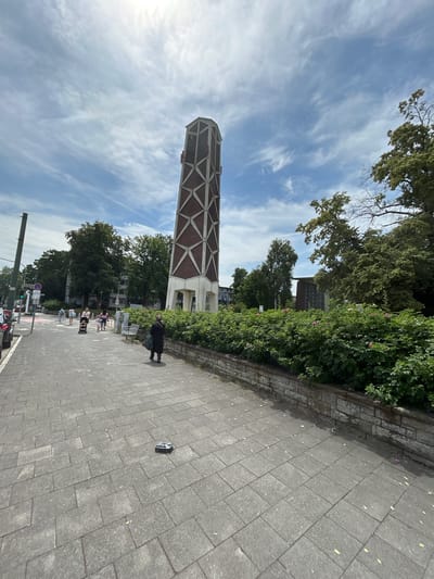 The image shows a tall, modern tower with a geometric pattern of brown and white material against a bright blue sky with wispy white clouds. The tower is situated next to a lush green hedge and a tree-lined street. A paved walkway runs alongside the hedge and street, and several people are walking on it. In the foreground, a person in dark clothing is standing near the hedge. Further down the walkway, a group of people, some appearing to be children, are gathered. Traffic lights and street signs are visible further down the street. On the walkway, a small, dark object, possibly a dropped item, is visible. The scene appears to be set during the daytime, under clear or partly cloudy skies. The presence of trees and people suggests an urban or suburban environment. The orientation of the image is vertical, with the tower dominating the left side and the walkway and street on the right.