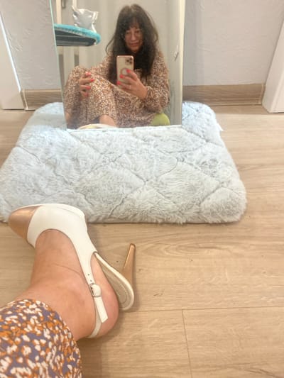 This media file shows a low-angle, full shot of a woman's feet and legs, as she takes a selfie in a mirror. The woman is wearing a floral dress and white and gold heeled sandals. The scene appears to be indoors, with a wooden floor and a fluffy, light-colored rug. In the mirror's reflection, the woman is visible, holding her phone and looking at the camera. She has dark hair and is wearing bright orange nail polish. The lighting suggests it could be daytime, and the setting looks like a domestic environment. There is no visible text in the image.