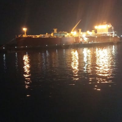 The image depicts a large industrial vessel docked at night in the city of Dock Sud, Argentina. The vessel features a dark red hull and is brightly illuminated by numerous artificial lights on its deck, revealing multiple tanks, extensive piping, and a visible crane. The letters "L P" are discernible on a mid-ship structure. The calm, dark water surface directly reflects these lights, creating prominent shimmering streaks of orange and yellow. No personnel, specific activities, or other vessels are clearly visible. The overall environment is dark, consistent with nighttime conditions.