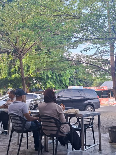 In Keuniree, Indonesia, a scene unfolds at an outdoor dining area adjacent to a road. Three individuals are visible, two seated at a metal table and one standing to the left. A man wearing a gray t-shirt and a blue and white striped cap is seated, facing away from the camera. He is wearing blue jeans. To his right, a person in a beige hijab and a light brown cardigan is also seated, facing away, with a small white purse on the table. A third individual, partially visible on the left, wears a striped shirt and is looking at a phone. On the table, there are some food items and a small white purse. In the background, a black SUV is parked on the road, along with several other vehicles. Behind the vehicles, a building with signage is visible, including advertisements for services like "DANA" and "GOPAY," and a sign that reads "IL PRANG POLEN." Trees with green foliage overhang the dining area, and a power line runs across the scene. The ground is paved with geometric tiles.