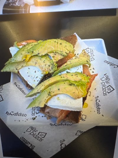 The image shows a close-up view of an open-faced sandwich with sliced avocado, cheese, and tomato, served on a white paper liner with logos that read "entre Plazas" and "Cafeteo". The sandwich appears to be on a slice of toast or bread. The setting is likely a cafe or restaurant. The "entre Plazas" logo suggests a location within a plaza or multiple plazas. Based on the prevalence of "Cafeteo" as a cafe chain in Spain, and the commonality of cafes within city centers, this could be a cafe in Murcia, Spain. There are no people visible in the image.