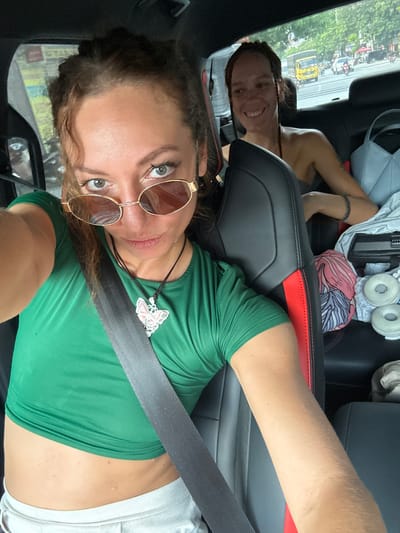 A selfie taken inside a car captures two women. In the foreground, a woman with light brown, curly hair, wearing amber-tinted sunglasses, looks directly at the camera with a calm expression. She wears a green crop top, exposing her midriff, and light-colored bottoms. A black seatbelt crosses her chest diagonally, and she has a butterfly pendant necklace. Her left arm is extended to take the photo, with her elbow resting on the car's black and red accented seat.
In the background, in the back seat, another woman with dark hair, possibly in dreadlocks or braids, smiles broadly at the camera. The car's interior features black leather seats with red detailing. Various items are visible on the back seat, including white headphones, a light grey bag, and a patterned fabric item.
Through the car window, a bright daytime street scene unfolds. Green trees line the road, and multiple vehicles, including motorcycles and a distinct yellow truck or bus, are visible, suggesting a busy thoroughfare. The setting is inside a moving or stopped vehicle, likely in Hà Giang 1 Ward, Vietnam. The time of day is clearly daytime, indicated by the bright outdoor lighting. No discernible text is visible.