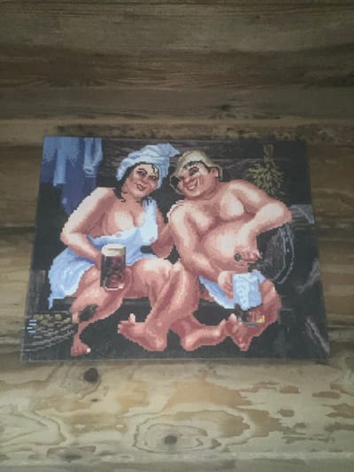 A pixel art image depicting two individuals in a banya-like setting is affixed to a light-brown wooden plank wall. The image features a woman on the left, with dark hair and a white towel wrapped around her head, wearing a light blue towel wrapped around her body, exposing her shoulders and chest. She holds a dark beverage in a glass mug and smiles toward the viewer. To her right is a stout man, wearing a light-colored hat and a light blue towel around his lower body. He holds a lighter beverage in a glass mug and a dark object in his other hand, also smiling toward the viewer. The background of the artwork shows a dark wooden interior, a barrel, a hanging *venik* (bath whisk), and blue towel-like items. The wooden wall exhibits prominent grain and knot patterns. The specific location within the city of Пугачёво, Russia, cannot be determined from the provided image.